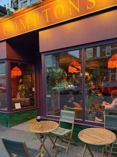 The Comptons shopfront at dusk, with an illuminated gold sign above deep aubergine woodwork, glowing amber lampshades in the window and pavement tables set out front.