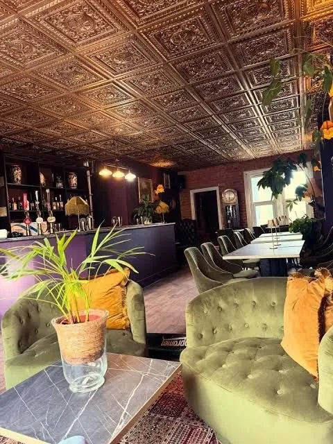The lounge corner at Comptons, with buttoned olive velvet tub chairs, a potted palm on a marble table and a patterned rug beneath the ornate gold ceiling.